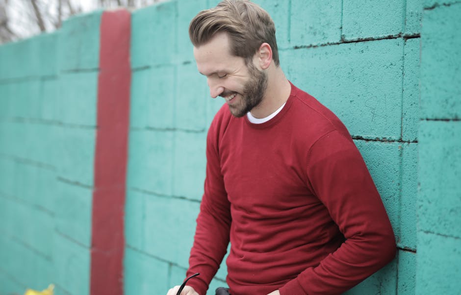 A man in a red sweater smiles while leaning against a colorful green wall outdoors.