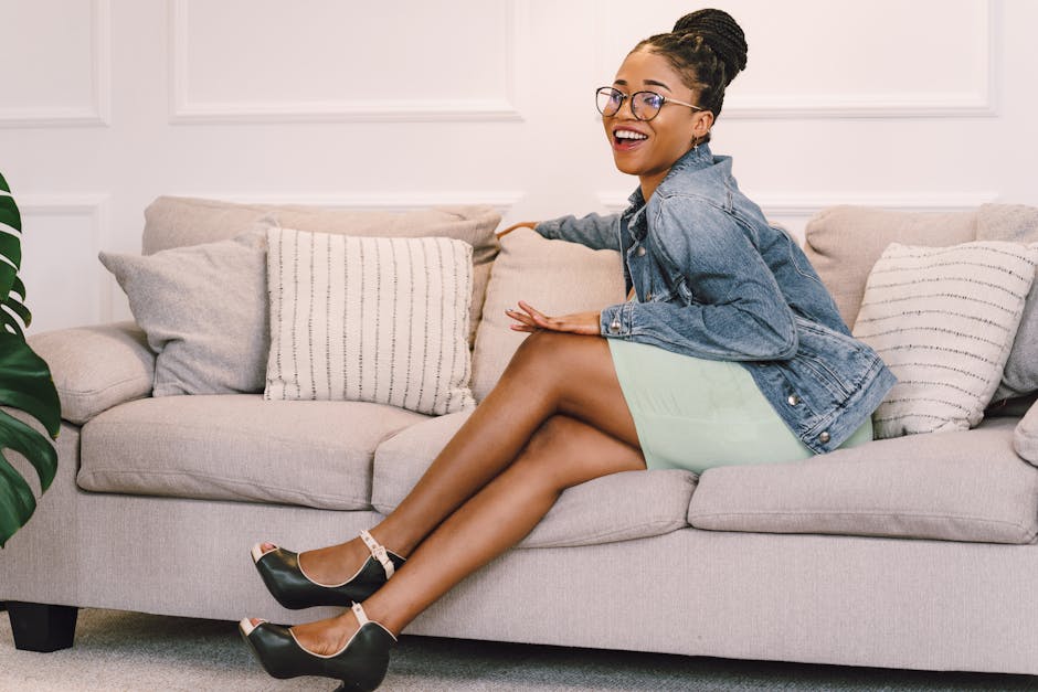 A cheerful woman in a denim jacket and mint dress smiles while sitting on a cozy sofa indoors.