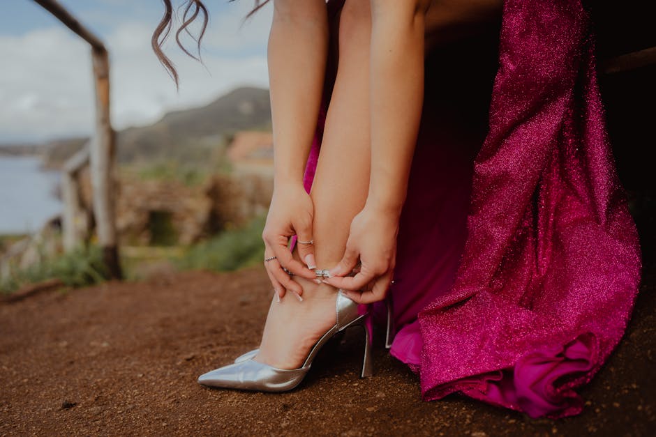 Elegant woman adjusting silver high heel with a sparkling pink gown in an outdoor setting.