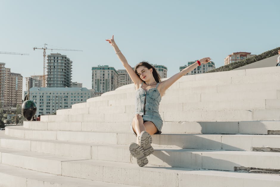Young woman in denim dress sitting on steps, arms raised joyously against a cityscape background.