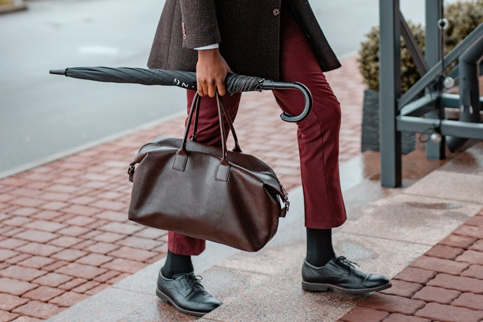 Man dressed in formal wear walking with a leather bag and umbrella on a rainy day.